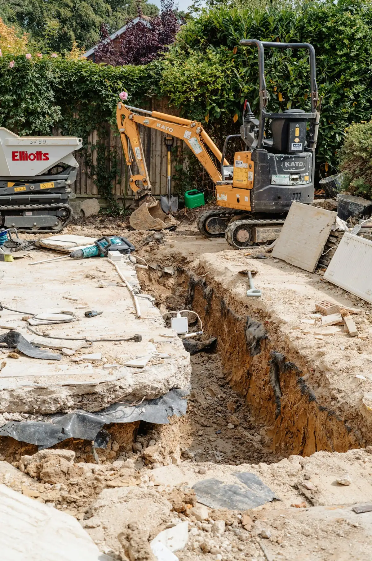 A foundation trench dug beside a house, with a mini digger and dumper behind